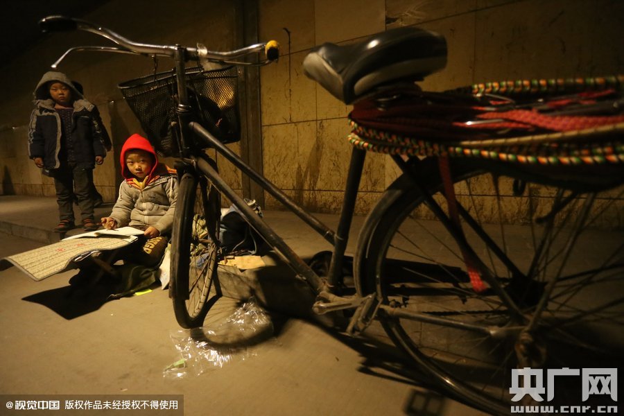 2015年11月8日晚，鄭州，7歲男孩涵洞橋下陪父母練攤，路邊燈下坐小馬扎上寫作業(yè)。